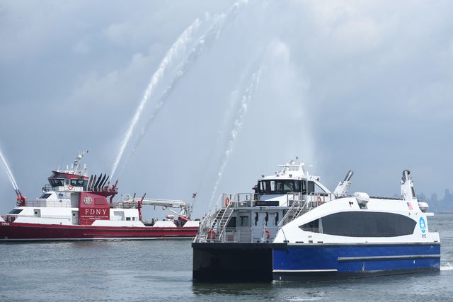 The new NYC ferry, which is blue and white, is in the water with an FDNY fire boat sending ceremonial streams of water